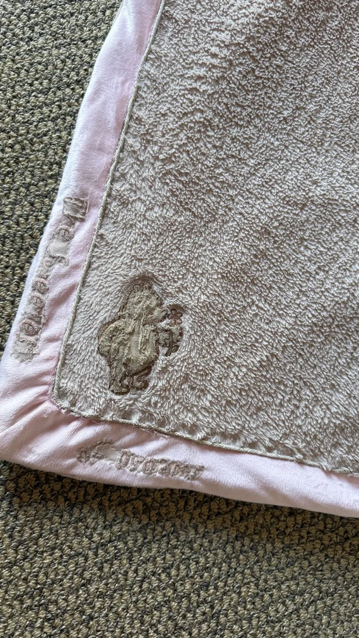 A well-loved pale blanket with pink trim and an embroidered teddy detail.