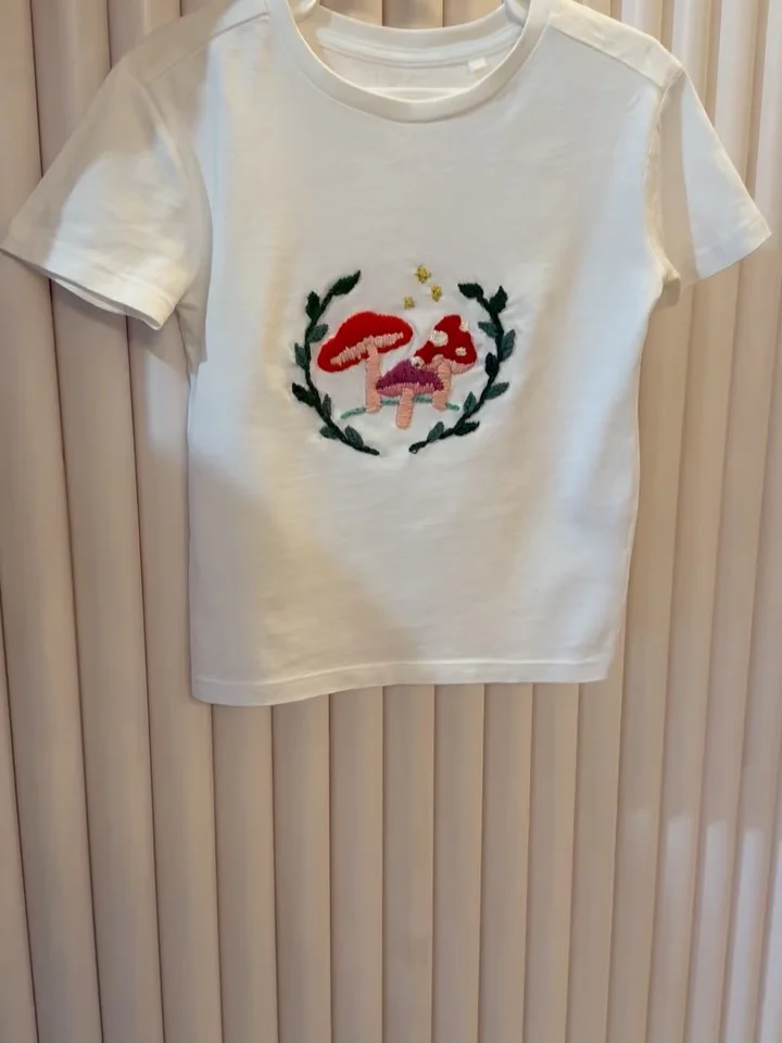 A white child’s T-shirt decorated with hand-embroidered red mushrooms and green foliage.